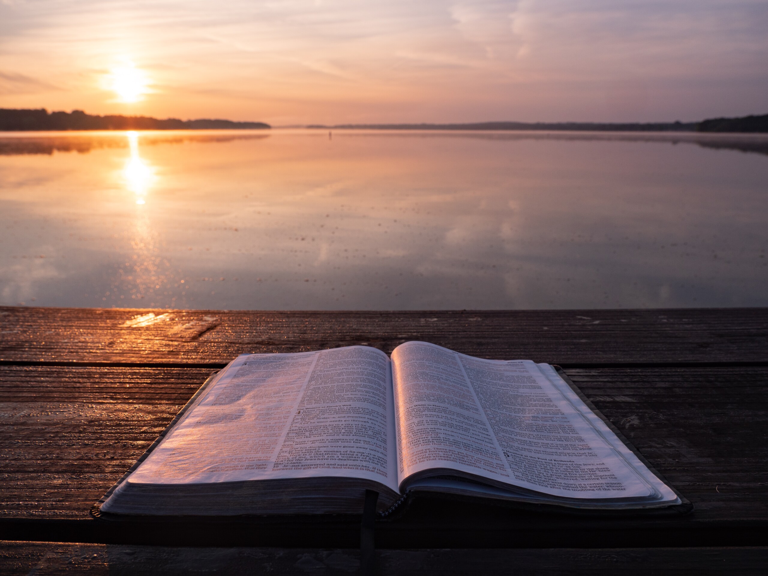 open bible on a table overlooking a body of water at sunrise/sunset